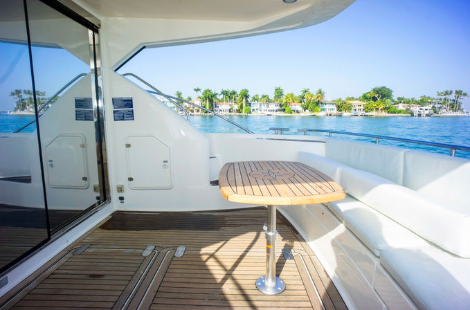 Sunlit luxury yacht deck with teak table and white cushioned seating, overlooking clear blue water and a palm-lined tropical waterfront with homes