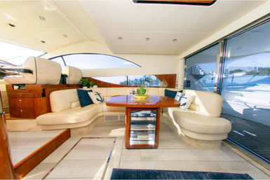 Bright luxury yacht salon with cream leather L-shaped seating, polished wood dining table, nautical anchor pillows and sliding glass doors opening to a sunny waterfront view.