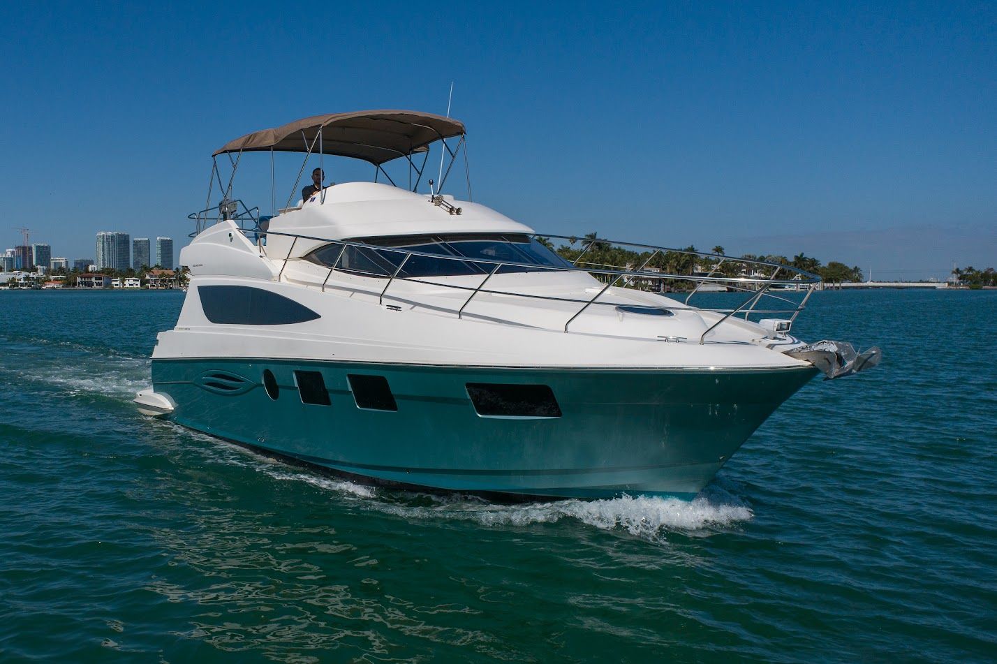 Sleek white motor yacht gliding through turquoise coastal waters past a palm-lined shore and distant city skyline under a clear blue sky.