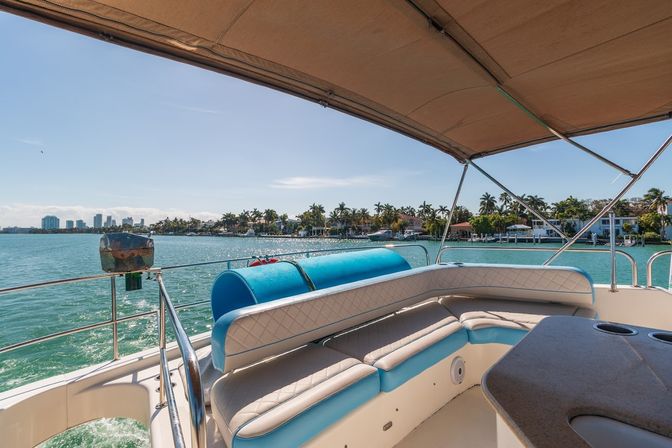 Sunlit boat deck with turquoise-and-white cushioned seating under a tan canopy, overlooking turquoise water, palm-lined waterfront homes and a distant city skyline under a clear blue sky
