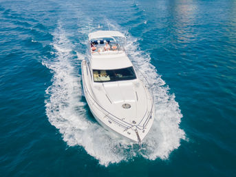 Aerial view of a luxury white motor yacht cruising on clear turquoise water, leaving a foamy wake with passengers relaxing on the sunlit upper deck.