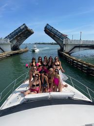 Group of women in bikinis smiling and holding pink cups on the bow of a white yacht passing under a raised drawbridge on a sunny coastal waterway with another boat in the channel and blue sky