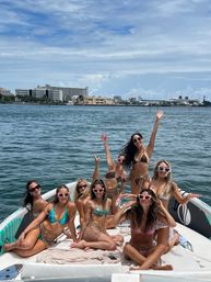 Eight women in colorful bikinis and heart-shaped sunglasses posing and cheering on a boat near an urban waterfront harbor under blue skies.