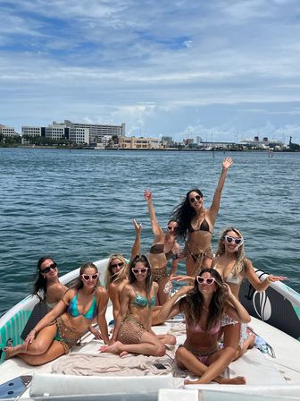 Eight women in colorful bikinis and heart-shaped sunglasses posing and cheering on a boat near an urban waterfront harbor under blue skies.