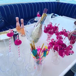 Flamingo-themed boat party table with champagne in an ice bucket, mini rosé bottles, pink paper flamingo straws, clear plastic flutes and colorful tropical stirrers, ocean visible in background.