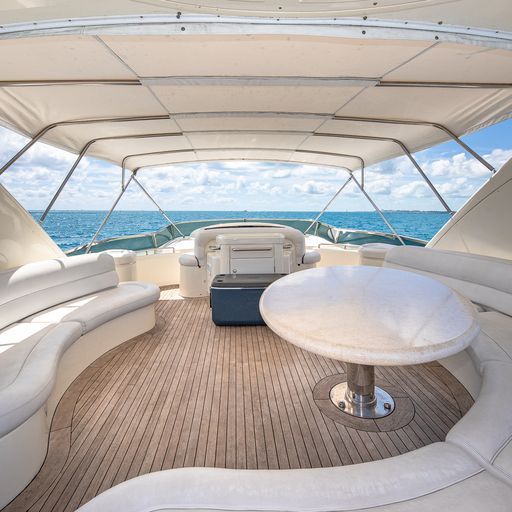 Sunlit luxury yacht aft deck with curved white seating, round table, teak deck and canvas bimini, overlooking turquoise open sea and blue sky.