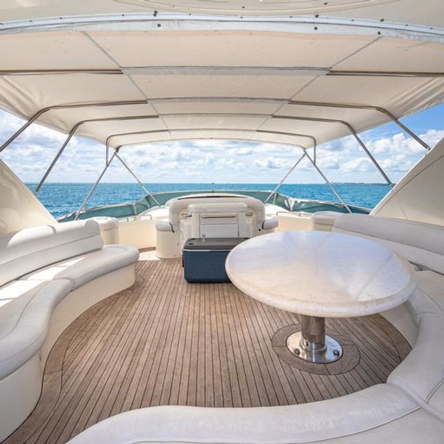 Sunlit luxury yacht aft deck with curved white seating, round table, teak deck and canvas bimini, overlooking turquoise open sea and blue sky.