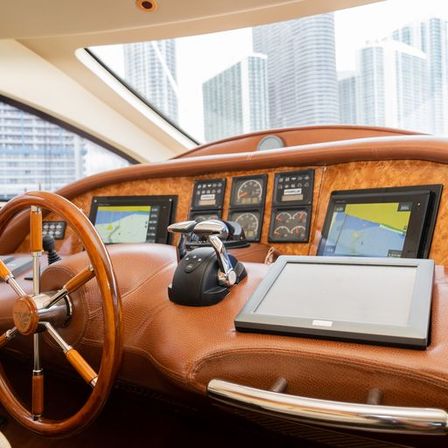 Sunlit luxury yacht helm with polished wooden steering wheel, leather-wrapped dashboard, twin marine GPS screens, chrome throttle controls and gauges, urban high-rise skyline visible through the windshield