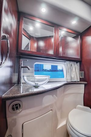 Compact yacht bathroom (boat head) with glass vessel sink, chrome faucet, dark wood mirrored cabinets, granite countertop and a porthole window framing a waterfront skyline.