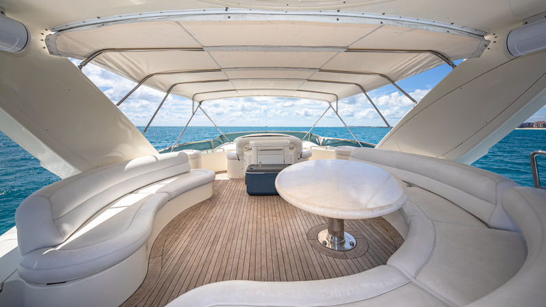 Sunlit covered yacht lounge with curved white leather seating and round table on teak deck, overlooking turquoise ocean and blue sky horizon.