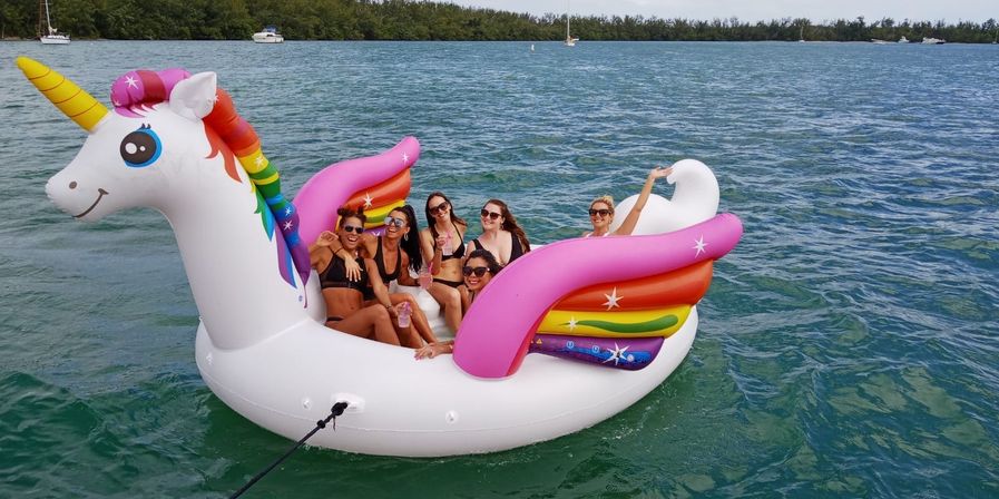 Six friends lounging on a giant inflatable rainbow unicorn float in turquoise coastal water, smiling and holding drinks with boats in the background