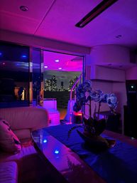 Luxury yacht interior at night with vibrant purple-blue LED lighting, orchid centerpiece on a glossy table, plush seating and an open deck revealing a glowing waterfront city skyline.