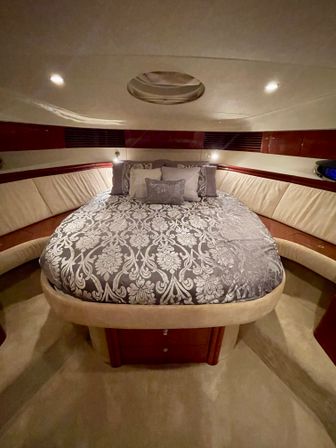 Luxury yacht forward stateroom with rounded queen bed dressed in gray damask bedding, plush beige leather wraparound seating, warm wood trim, overhead hatch and recessed lighting