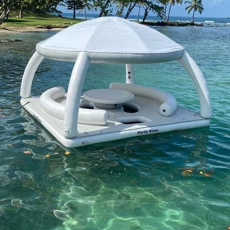 White inflatable floating lounge with canopy, circular seating and center table drifting on clear turquoise tropical water near a palm-lined shore.