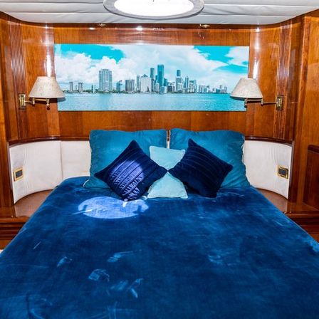 Luxury yacht cabin bedroom with deep-blue velvet bedding and teal/navy pillows, warm wood paneling and a panoramic coastal city skyline print above the bed.