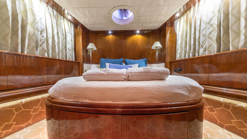 Cozy luxury yacht stateroom interior with rounded queen bed, glossy wood paneling, porthole skylight, bedside lamps and neutral curtains.