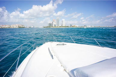 Bow of a white yacht cruising on turquoise ocean toward a sunny coastal city skyline with beachfront high-rise buildings and fluffy clouds