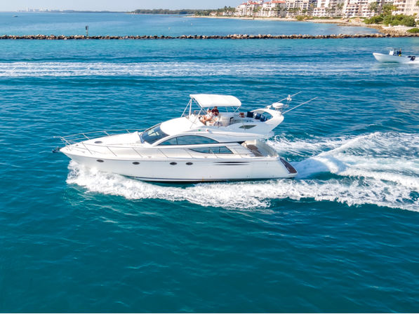 White luxury motor yacht cruising turquoise ocean on a sunny day, leaving a foamy wake with beachfront condos on the horizon and people relaxing on the upper deck