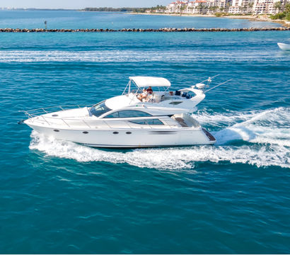 White luxury motor yacht cruising turquoise ocean on a sunny day, leaving a foamy wake with beachfront condos on the horizon and people relaxing on the upper deck