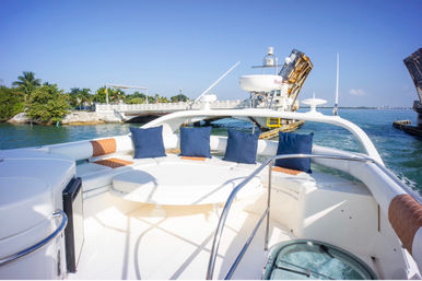 Sunny luxury yacht bow lounge with white round table and navy pillows, cruising through a tropical channel under an open drawbridge with palm-lined shore and clear blue sky.