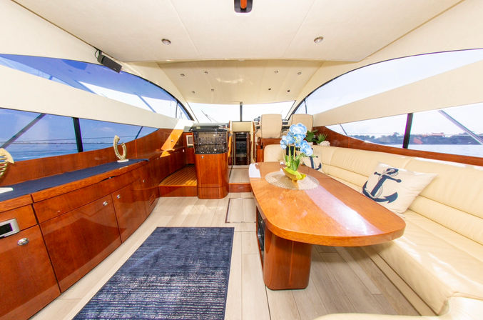 Sunlit luxury yacht salon with glossy wood cabinetry, cream leather L‑shaped sofa, wooden dining table crowned by blue flowers, nautical anchor pillows and panoramic windows framing an ocean view.