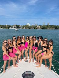 Group of women in black swimsuits and pink sarongs posing on the bow of a yacht under a sunny blue sky with palm-lined shoreline and city skyline on the Miami waterfront.