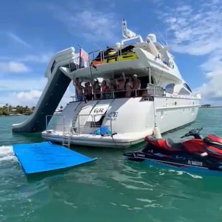 Luxury white yacht anchored in turquoise coastal water with a large inflatable water slide off the stern, people gathered on the aft deck, a blue floating mat and a red-and-black jet ski alongside under a sunny sky