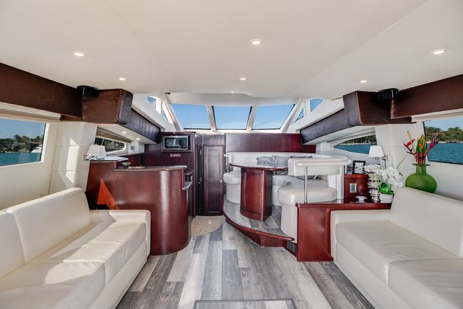 Sunlit luxury yacht salon interior with white leather sofas, curved dark-wood bar and raised dinette, panoramic windows and skylights showing blue coastal water, gray wood-look flooring and decorative plants.