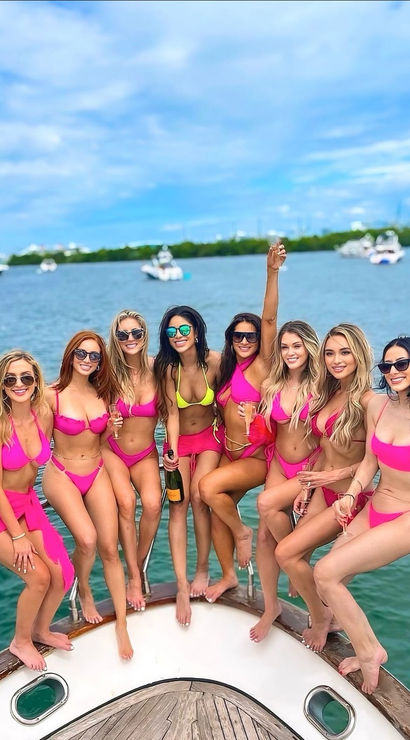 Eight women in bright pink bikinis posing and cheering with champagne on the bow of a yacht, turquoise coastal water and anchored boats under a bright blue sky — celebratory summer boat day vibe.