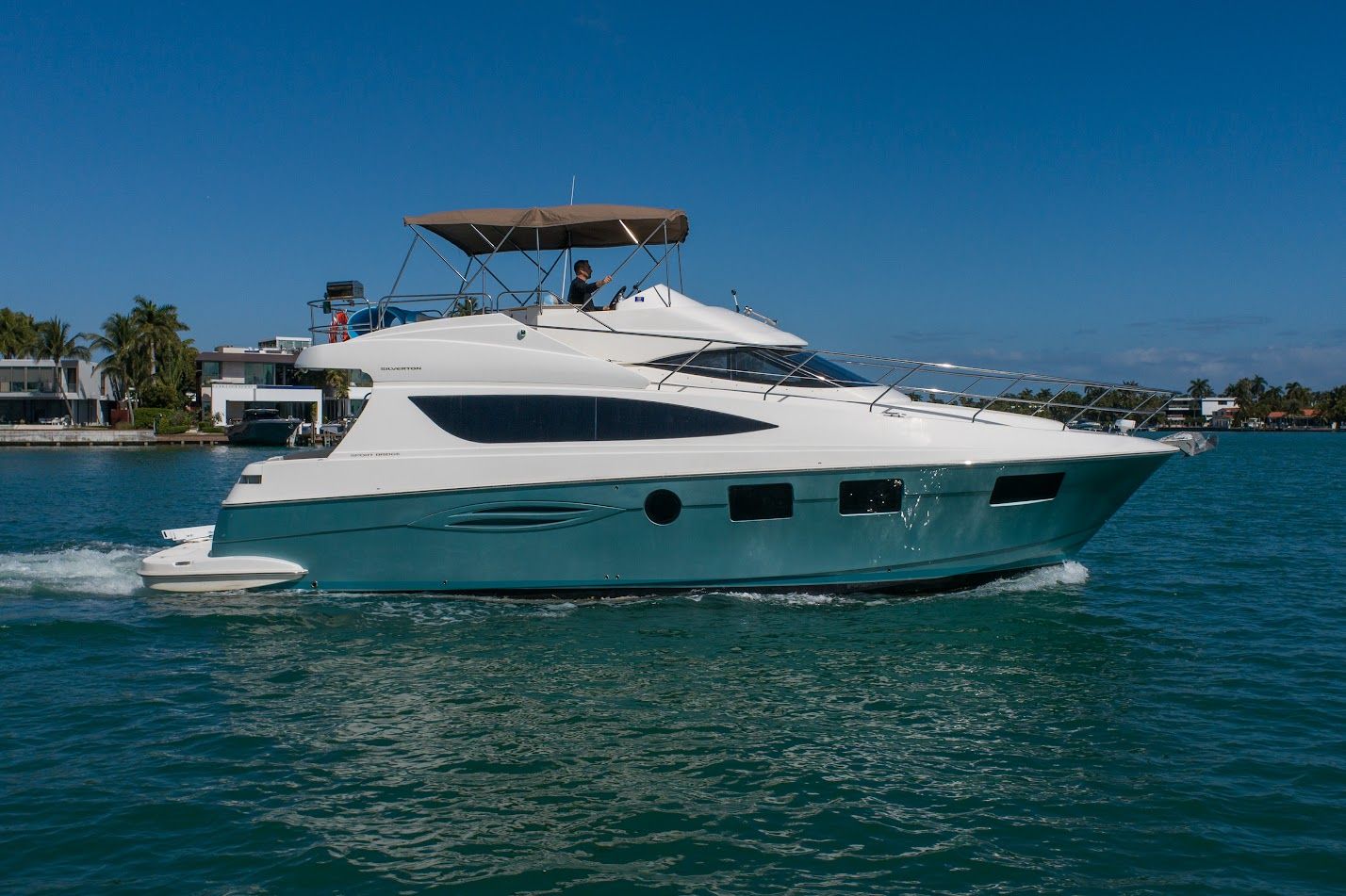 Teal-and-white luxury motor yacht cruising past waterfront homes and palm trees on a sunny blue-water coastal channel