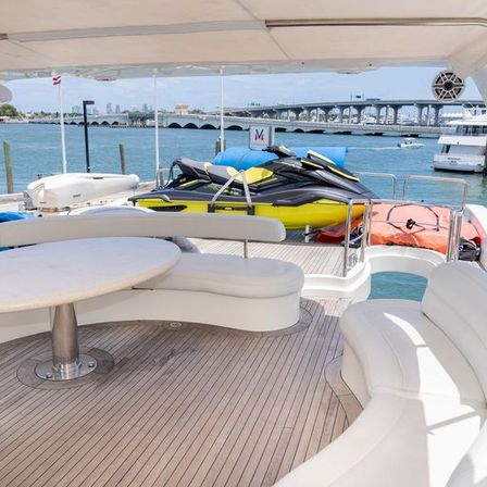 Luxury yacht deck with curved white seating and table, bright yellow-and-black jet skis on the stern, marina waters and arched bridge with city skyline in the background