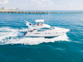 Luxury white motor yacht slicing through turquoise water, leaving a foamy wake near a rocky breakwater and marina pier with a coastal skyline in the distance on a sunny day