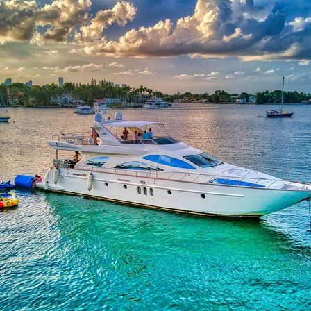 Luxury white yacht anchored in turquoise coastal bay at sunset, people on deck with inflatable tubes tied to the stern, palm-lined shoreline and other boats under dramatic clouds
