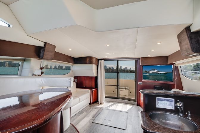 Sunlit luxury yacht interior salon with white leather seating, glossy wood table, compact galley sink and sliding glass doors opening to blue water and a coastal city skyline.