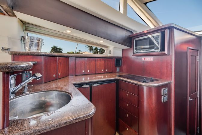 Compact yacht galley with rich cherry wood cabinets, granite-look countertops, stainless sink, microwave, two-burner cooktop and mini-fridge, bright port window showing palm trees and blue sky for a coastal vibe.
