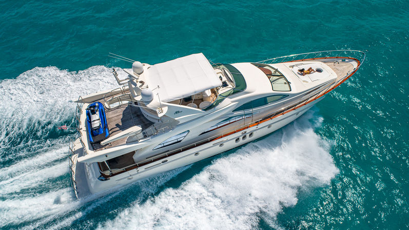 Aerial view of a white luxury motor yacht cutting through clear turquoise sea, leaving a white wake, with a blue jet ski on the aft deck and a person sunbathing on the bow.