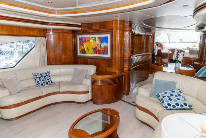 Sunlit luxury yacht salon with curved cream leather sofas, glossy wood paneling, glass coffee table, dining area and marina view through large windows