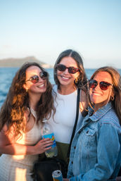 Three friends in sunglasses smiling on a sunset boat cruise, holding drinks with ocean and distant coastline in the background.