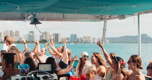 Group of friends partying on a boat in turquoise water with the Honolulu Waikiki skyline and Diamond Head crater visible on the horizon