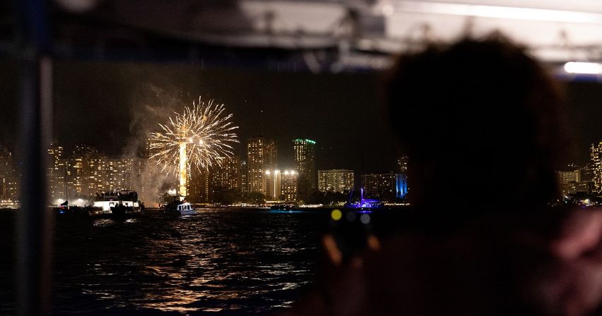 Nighttime fireworks burst over a lit city skyline and harbor, boats on the water and a silhouetted spectator watching from a nearby boat.