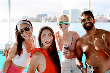 Four friends smiling on a sunlit boat with a waterfront city skyline behind them, wearing summer outfits and sunglasses, holding canned drinks and enjoying a coastal day out
