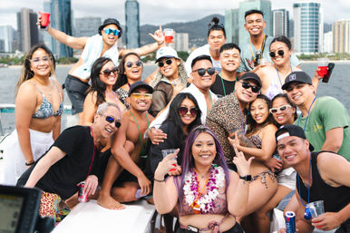 Large group of smiling friends at a lively boat party on the water with a high-rise city skyline in the background, many wearing swimsuits, leis and sunglasses and holding drinks.