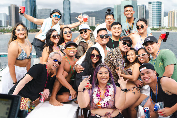 Smiling group of friends on a sunny boat party off a coastal city skyline, wearing swimsuits, leis and sunglasses, holding drinks and posing for a photo