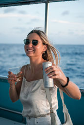 Smiling woman in sunglasses on a boat deck holding a canned drink and plastic cup with the open ocean as a backdrop — relaxed cruise vibe.