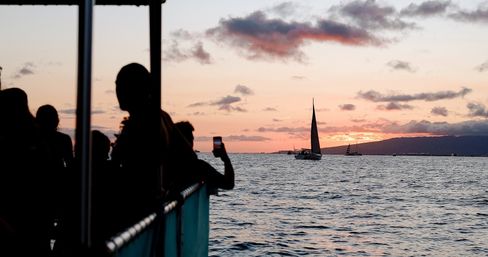 Silhouetted ferry passengers watch sailboats glide across calm ocean waters at a pink-orange sunset with an island silhouette on the horizon.