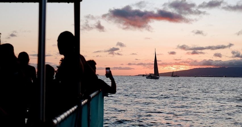 Silhouetted ferry passengers watch sailboats glide across calm ocean waters at a pink-orange sunset with an island silhouette on the horizon.