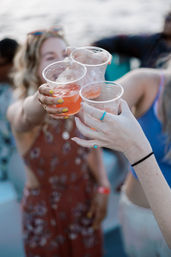 Three friends clinking plastic cups of pink iced cocktails at a sunny waterfront summer party
