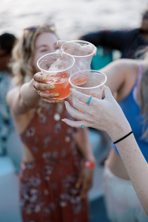 Three friends clinking plastic cups of pink iced cocktails at a sunny waterfront summer party