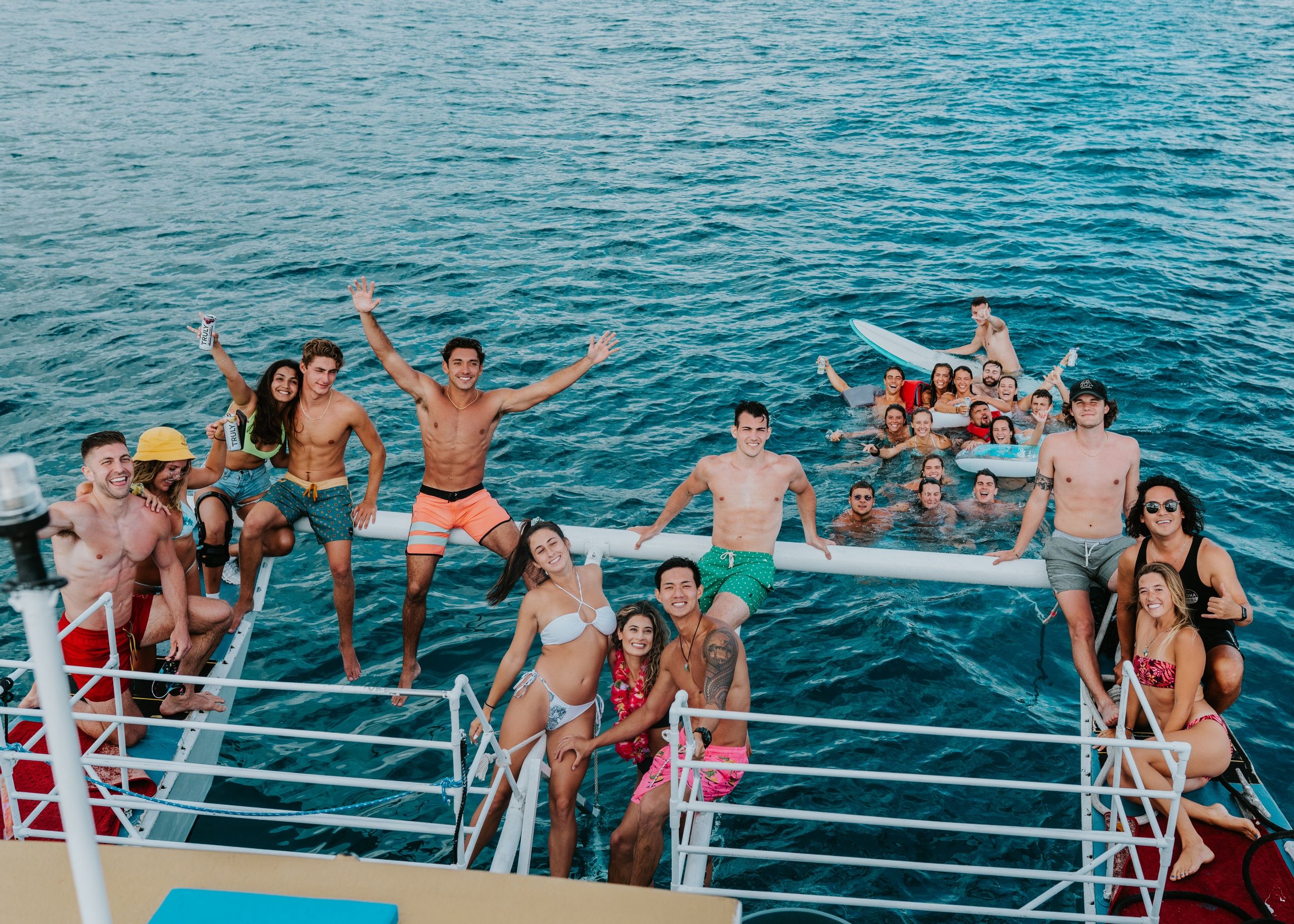 Boat party with a large group of friends in swimsuits enjoying clear turquoise ocean—people on the boat’s outriggers and floating nearby with surfboards and drinks