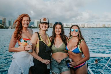 Four smiling women in colorful swimsuits and summer outfits posing on a boat with blue ocean and a coastal city skyline in the background, holding canned drinks and enjoying a sunny day